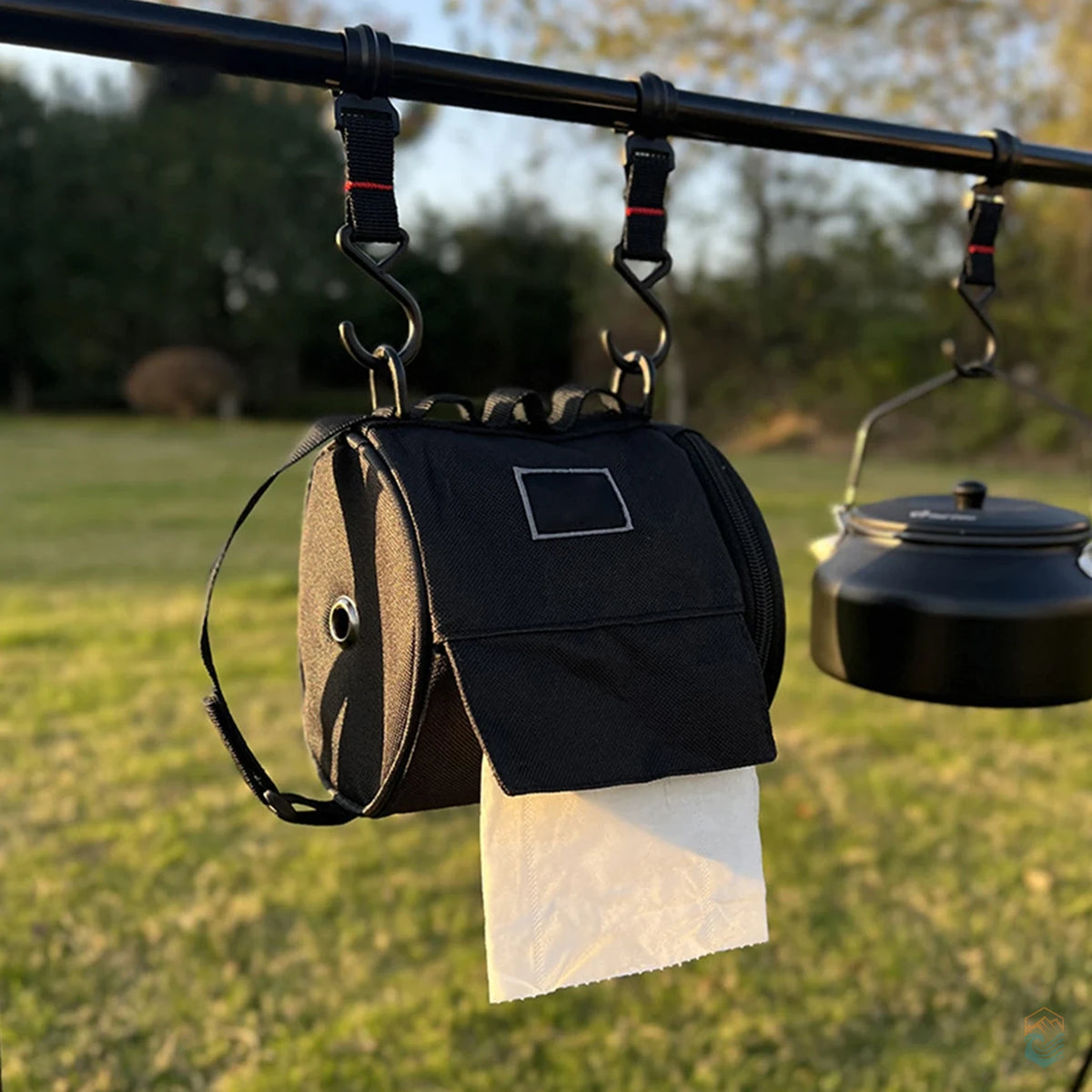Black outdoor toilet paper storage bag hanging from a camping rack next to a kettle, dispensing tissue in a grassy campsite setting