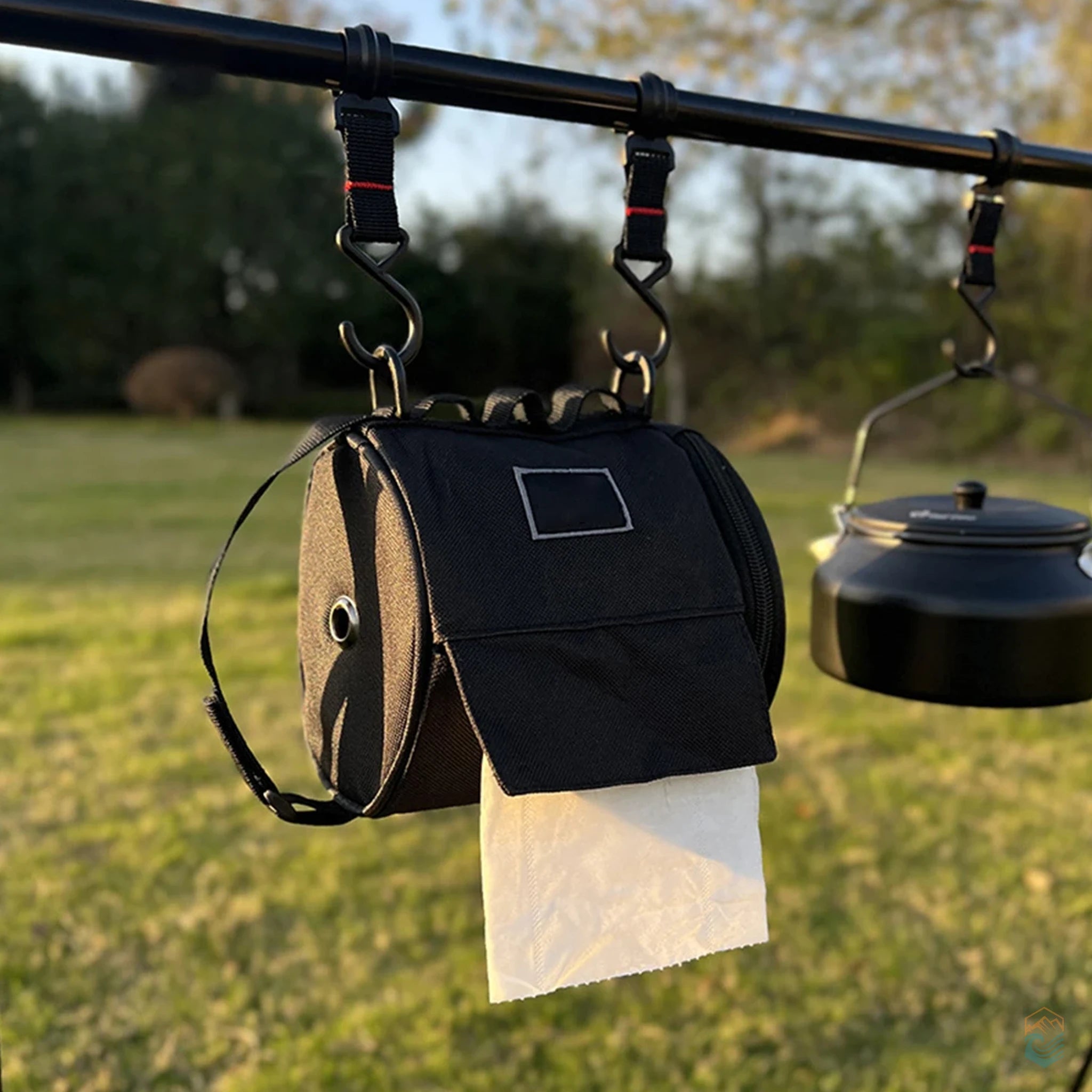 Black outdoor toilet paper storage bag hanging from a camping rack next to a kettle, dispensing tissue in a grassy campsite setting