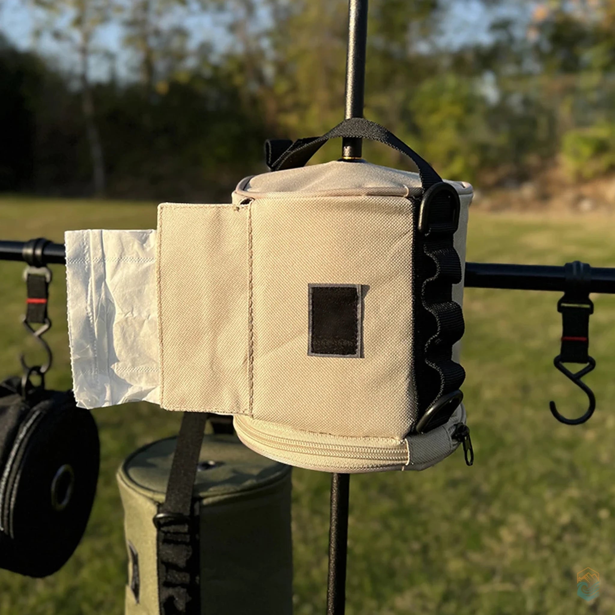 Khaki outdoor toilet paper storage bag hanging from a camp rack, with tissue pulled through the front flap and side loops visible in a grassy outdoor setting