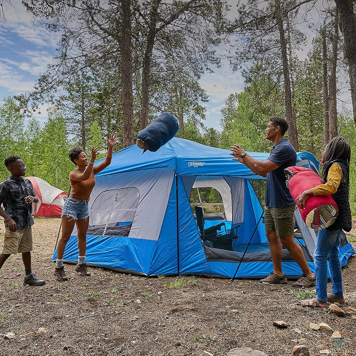 Family enjoying a camping trip beside the Wenzel Klondike 8-person tent in the forest, tossing sleeping bags and setting up camp