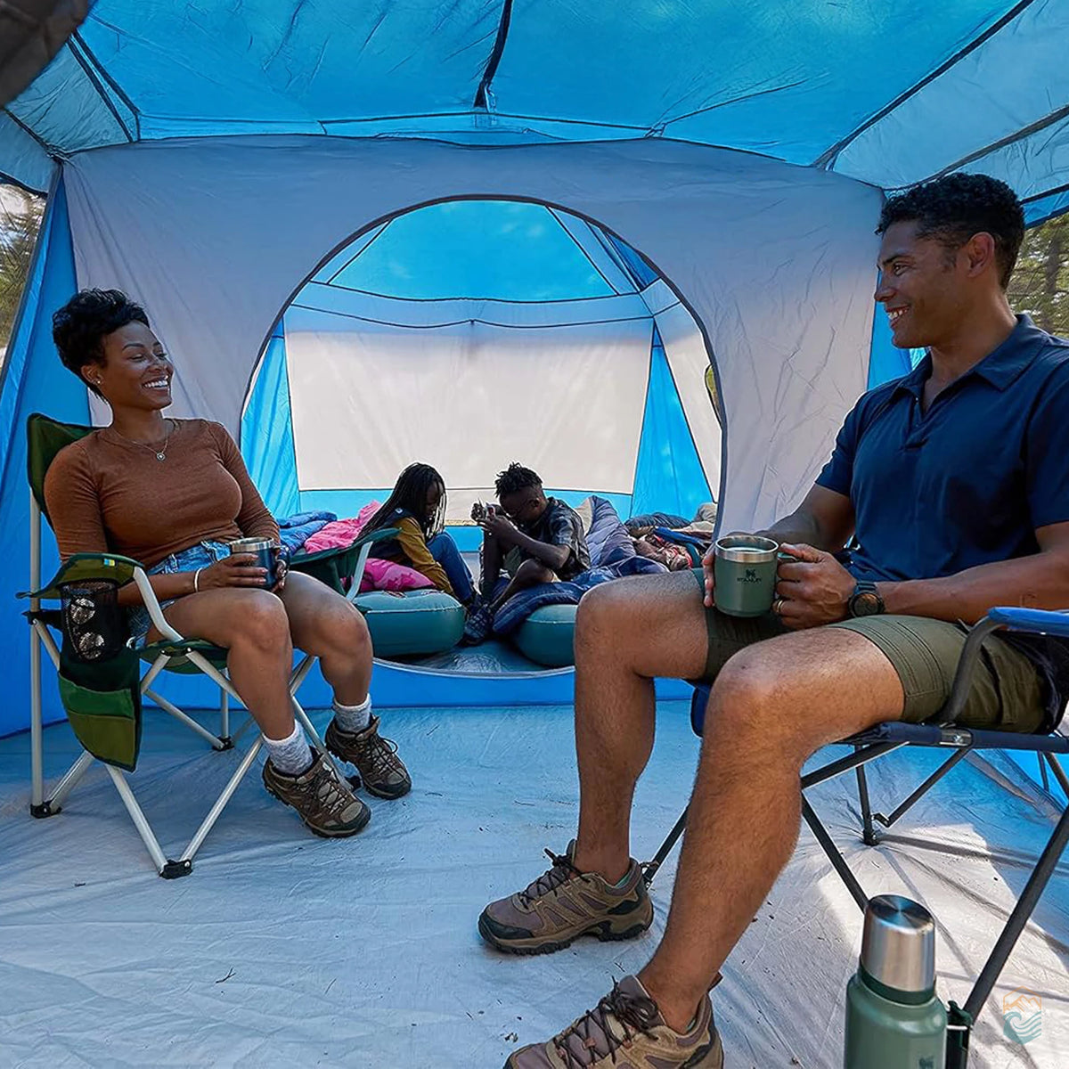 Family relaxing inside the Wenzel Klondike 8-person tent, parents seated with coffee while kids play on air mattresses in the back room