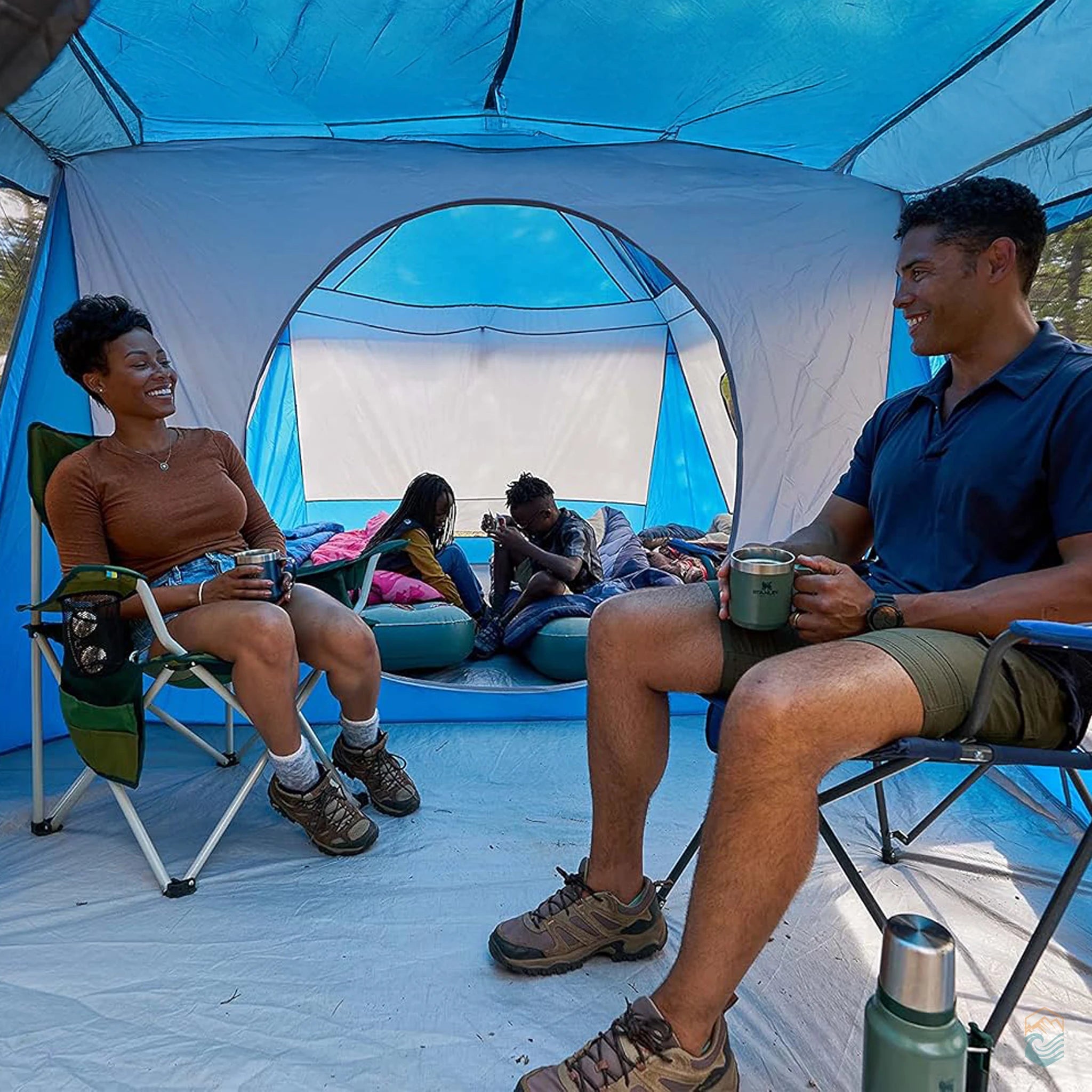 Family relaxing inside the Wenzel Klondike 8-person tent, parents seated with coffee while kids play on air mattresses in the back room