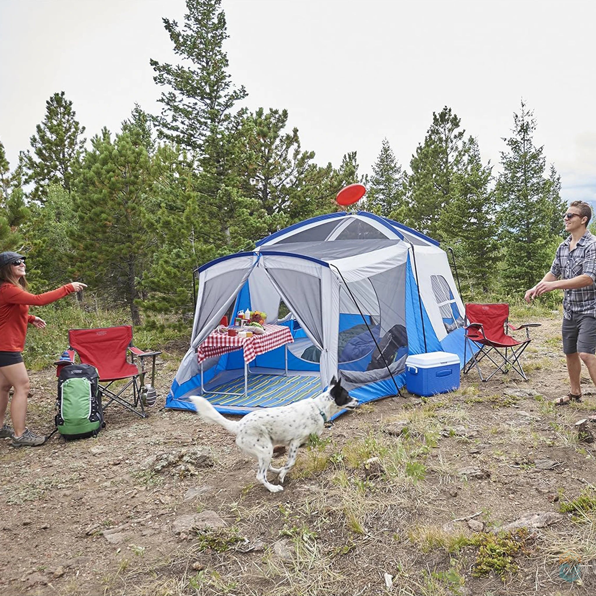 Couple playing frisbee with their dog in front of a Wenzel Klondike tent with a screen room and picnic setup during a forest camping trip