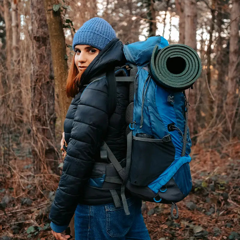 A woman wearing a blue beanie and black jacket hiking in a forest, carrying a blue backpack with a rolled-up sleeping pad.