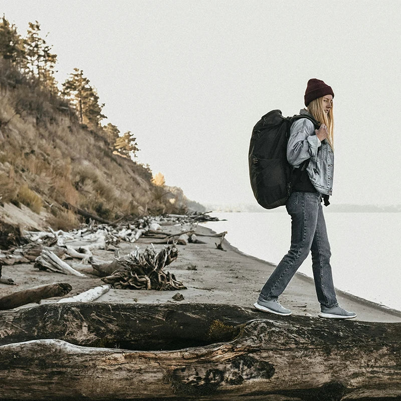 A woman hiking along a beach, carrying a large backpack and walking on a log, with driftwood and cliffs in the background.