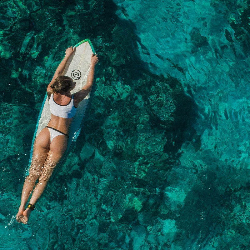 A woman on a surf board paddling over a crystal clear water, seen from above, showcasing the beautiful underwater landscape.