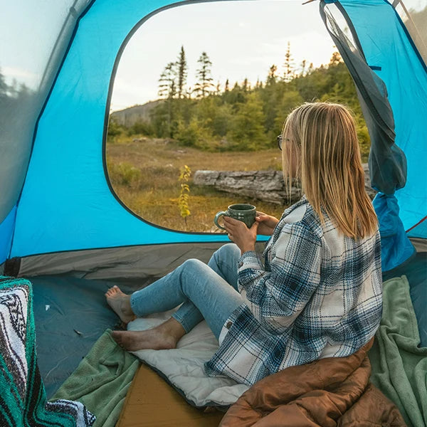 A woman sitting inside a blue camping tent, sitting on sleeping bags, holding a cup of coffee and enjoying the view of a grassy field and trees outside.